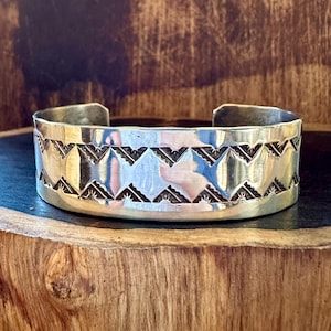 May include: A silver cuff bracelet with a geometric pattern. The bracelet has a wide band with a series of repeating shapes. The design is etched into the metal, creating a contrast between the shiny silver and the darker recessed areas.