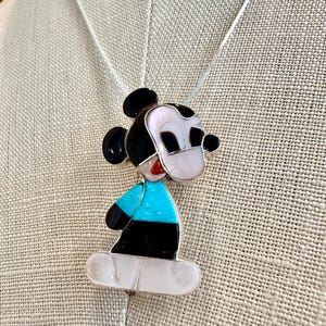 K&ouml;nnte beinhalten: Eine Halskette mit Mickey Mouse Anh&auml;nger. Der Anh&auml;nger hat ein farbenfrohes Design mit schwarzen, wei&szlig;en, roten und t&uuml;rkisblauen Akzenten. Die Halskette hat eine silberne Kette.