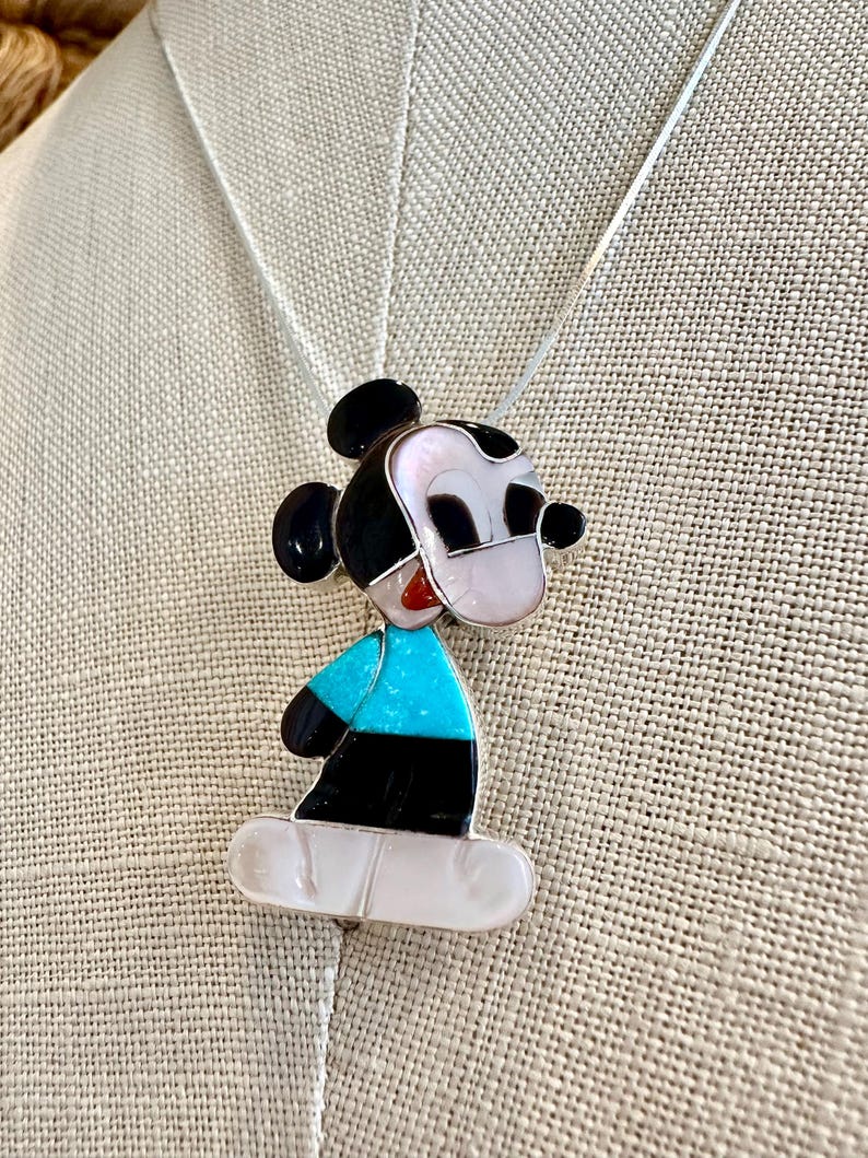K&ouml;nnte beinhalten: Eine Halskette mit Mickey Mouse Anh&auml;nger. Der Anh&auml;nger hat ein farbenfrohes Design mit schwarzen, wei&szlig;en, roten und t&uuml;rkisblauen Akzenten. Die Halskette hat eine silberne Kette.