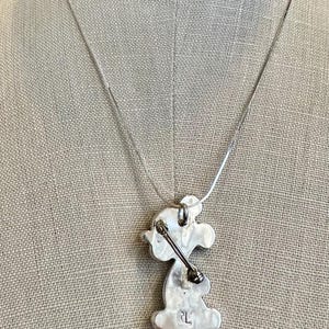 K&ouml;nnte beinhalten: Eine silberfarbene Halskette mit Anh&auml;nger, die eine stilisierte Figur zeigt. Der Anh&auml;nger hat eine einzigartige Form mit einem stiftartigen Element. Die Halskette wird auf einer Schaufensterpuppe pr&auml;sentiert. Das Wort "FORM" ist oben in blauen Buchstaben zu sehen.
