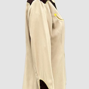 May include: A long-sleeved button-down shirt in beige with dark brown shoulder yokes and cuffs. The shirt features bright yellow piping along the yokes, cuffs, and a single chest pocket. The cuffs are elongated and secured with a row of small brown buttons.
