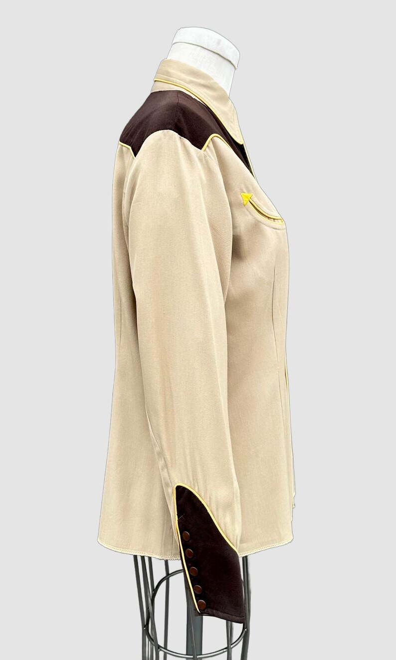 May include: A long-sleeved button-down shirt in beige with dark brown shoulder yokes and cuffs. The shirt features bright yellow piping along the yokes, cuffs, and a single chest pocket. The cuffs are elongated and secured with a row of small brown buttons.