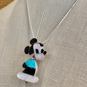 K&ouml;nnte beinhalten: Halskette mit Mickey Mouse Anh&auml;nger. Der Anh&auml;nger ist mit schwarzen, wei&szlig;en, t&uuml;rkisfarbenen und roten Akzenten versehen. Die Kette hat eine silberne Kette und wird auf einem neutralen Hintergrund pr&auml;sentiert.