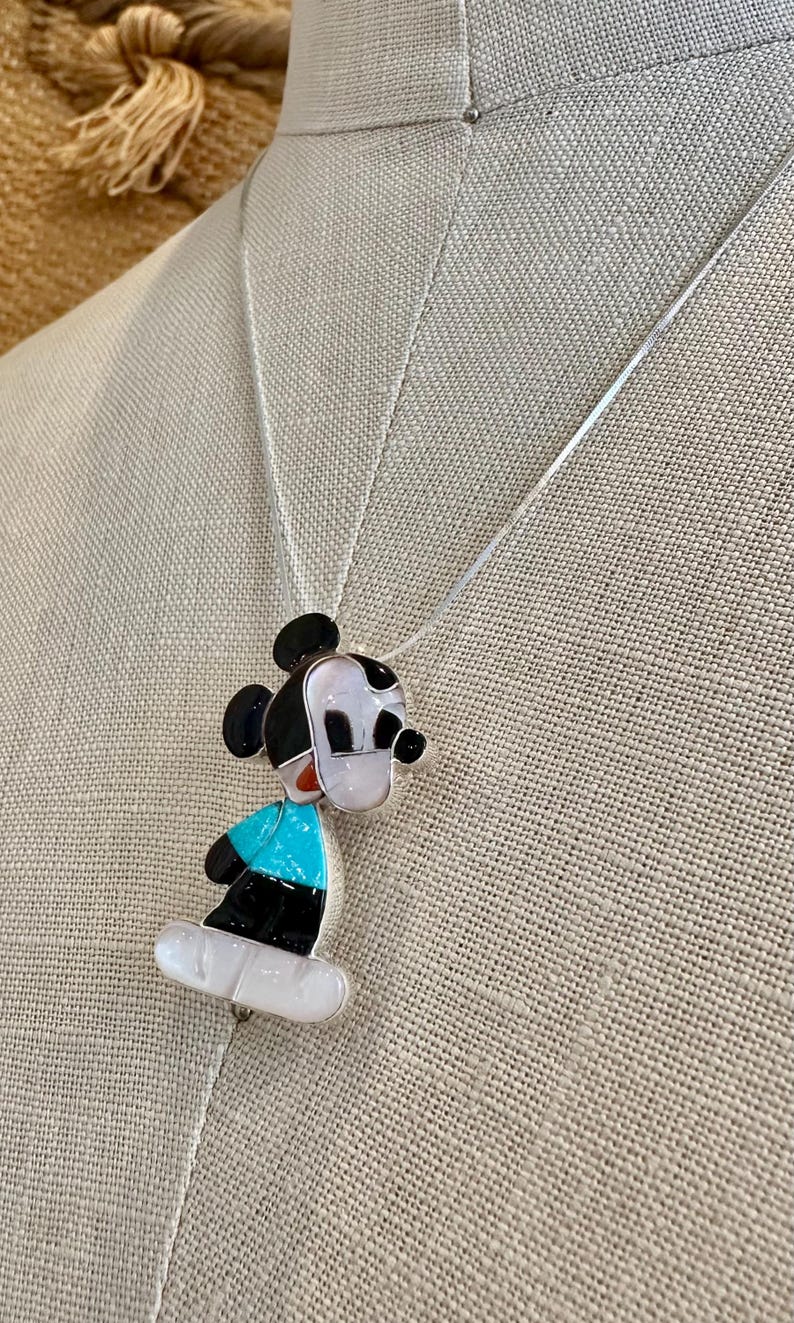 K&ouml;nnte beinhalten: Halskette mit Mickey Mouse Anh&auml;nger. Der Anh&auml;nger ist mit schwarzen, wei&szlig;en, t&uuml;rkisfarbenen und roten Akzenten versehen. Die Kette hat eine silberne Kette und wird auf einem neutralen Hintergrund pr&auml;sentiert.