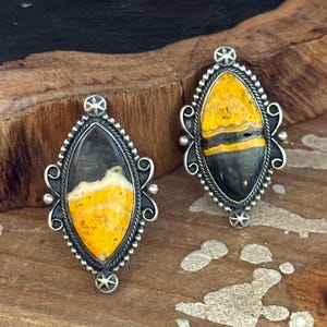 May include: Two marquise-shaped rings featuring bumblebee jasper stones with distinct yellow, black, and grey bands. Each stone is set in a decorative sterling silver frame with beaded edges, scrollwork accents, and a small star detail at the top and bottom.