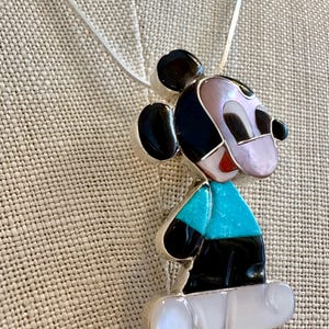 K&ouml;nnte beinhalten: Eine Halskette mit einem Mickey Mouse-Anh&auml;nger. Der Anh&auml;nger hat ein farbenfrohes Mosaikdesign mit schwarzen, t&uuml;rkisfarbenen, wei&szlig;en und rosa Elementen. Die Halskette hat eine silberne Kette.