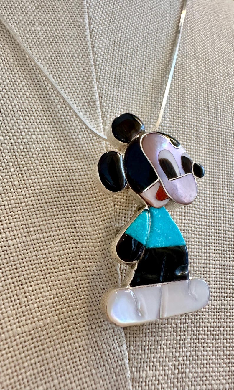 K&ouml;nnte beinhalten: Eine Halskette mit einem Mickey Mouse-Anh&auml;nger. Der Anh&auml;nger hat ein farbenfrohes Mosaikdesign mit schwarzen, t&uuml;rkisfarbenen, wei&szlig;en und rosa Elementen. Die Halskette hat eine silberne Kette.