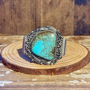May include: A silver cuff bracelet featuring a large, irregular, polished turquoise stone with brown matrix. The metal setting is decorated with intricate silver wire scrolls, small silver beads, and etched leaf-like patterns along the edges.