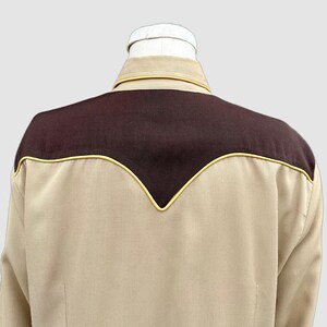 May include: A long-sleeved shirt in a light tan colour featuring a dark brown yoke across the upper back and shoulders. The yoke is accented with a thin yellow piping trim that follows the V-shaped contour of the seam, and the shirt has a collar.