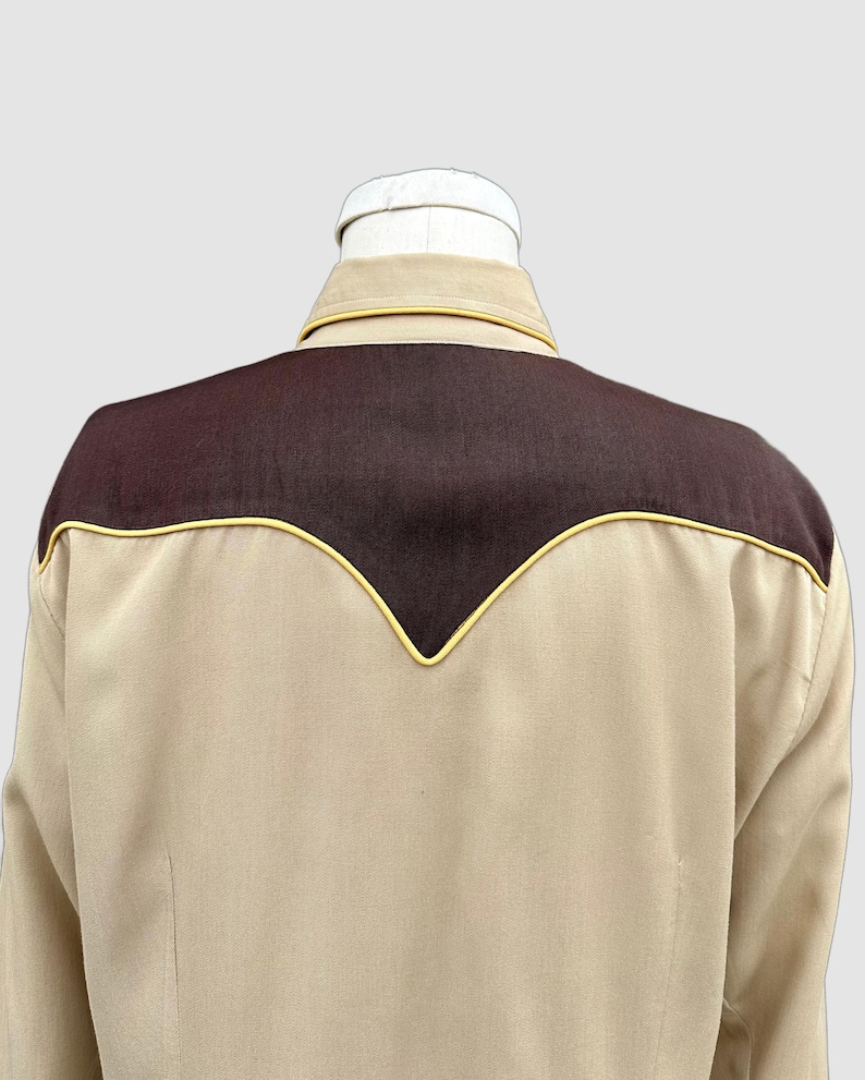 May include: A long-sleeved shirt in a light tan colour featuring a dark brown yoke across the upper back and shoulders. The yoke is accented with a thin yellow piping trim that follows the V-shaped contour of the seam, and the shirt has a collar.