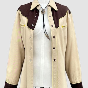 May include: A long-sleeve button-down shirt in beige and dark brown with a pointed collar and western-style yokes. The shirt features dark brown cuffs, yellow piping accents, and a bolo tie with a central oval pendant featuring a horse motif.