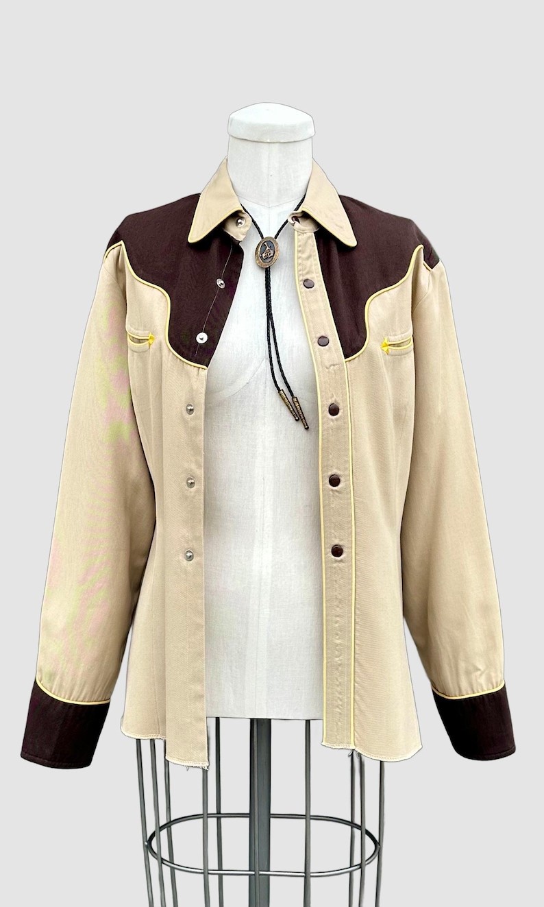 May include: A long-sleeve button-down shirt in beige and dark brown with a pointed collar and western-style yokes. The shirt features dark brown cuffs, yellow piping accents, and a bolo tie with a central oval pendant featuring a horse motif.