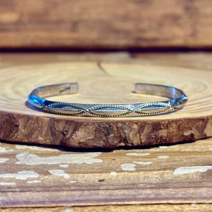 Puede incluir: Una pulsera de plata con un patrón de diamantes repetido. La pulsera está abierta y descansa sobre una superficie de madera. La pulsera tiene un acabado pulido y está ligeramente curvada.