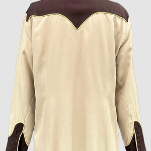 May include: A long-sleeved button-down shirt in beige with dark brown panels on the shoulders and cuffs. The shirt features a pointed yoke design on the back and sleeves, accented with thin yellow piping along the seams and brown buttons.