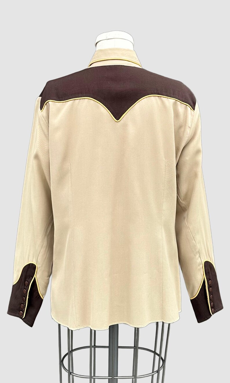 May include: A long-sleeved button-down shirt in beige with dark brown panels on the shoulders and cuffs. The shirt features a pointed yoke design on the back and sleeves, accented with thin yellow piping along the seams and brown buttons.