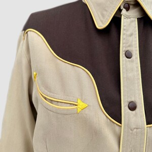 May include: A long-sleeve shirt featuring a tan body with a dark brown yoke across the chest and shoulders. The shirt includes yellow piping along the yoke, collar, and pocket, with brown snap buttons and a decorative yellow arrow-shaped embroidery.