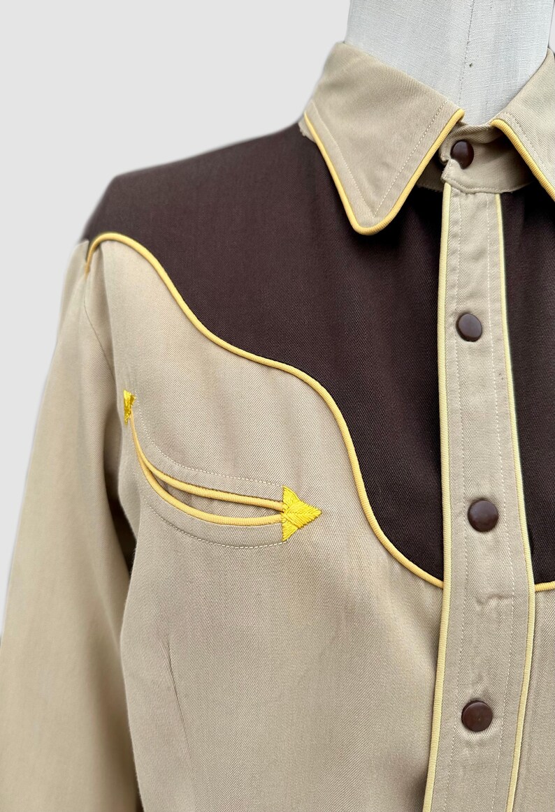 May include: A long-sleeve shirt featuring a tan body with a dark brown yoke across the chest and shoulders. The shirt includes yellow piping along the yoke, collar, and pocket, with brown snap buttons and a decorative yellow arrow-shaped embroidery.
