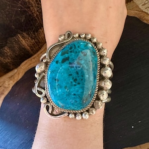 May include: A silver cuff bracelet featuring a large, oval turquoise stone. The stone is a vibrant teal color and is surrounded by a silver rope-like border. The bracelet has a detailed, ornate design with silver accents and small silver beads.