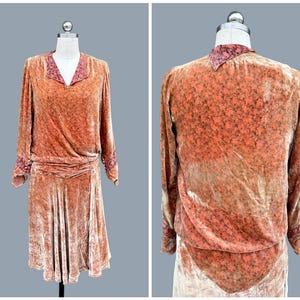 May include: A vintage peach-colored velvet dress with long sleeves and a V-neck collar. The dress features a floral pattern and a gathered waist. The cuffs and collar are accented with a darker floral fabric. The dress is displayed on a mannequin.