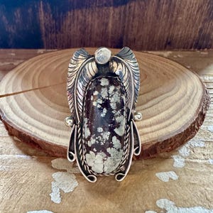 May include: A silver-toned ring with a large, oval-shaped stone. The stone is a mix of dark brown and cream colors. The ring has a detailed, feather-like design around the stone and a small, round accent stone at the top.