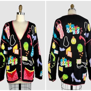 Può includere: Un cardigan nero con scollo a V caratterizzato da un motivo lavorato a maglia con borse della spesa, pacchi regalo, scarpe con tacco e cornette telefoniche. Il capo presenta un bordo rosa lungo l'apertura e due tasche con bottoni dorati.
