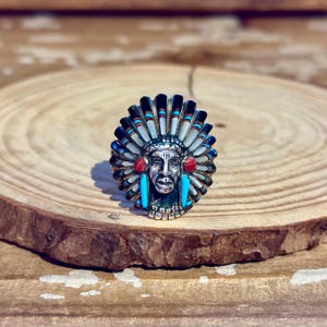 May include: A silver ring featuring a central face sculpture wearing a detailed headdress. The headdress is inlaid with black, white, red, and turquoise stone segments arranged in a radial pattern, with two red circular stones at the ears.