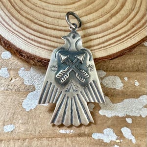 May include: A silver-coloured bird-shaped pendant with crossed arrows. The pendant has detailed engravings, including feathers, sunbursts, and arrowheads. It has a loop at the top for hanging, likely a piece of jewellery.
