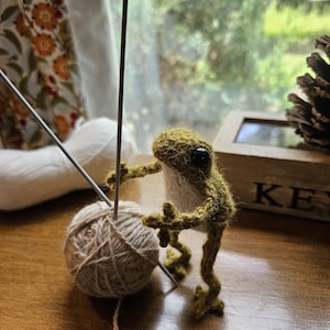 May include: A handmade, green crocheted frog figurine with a ball of yarn and knitting needles. The frog is positioned as if it is knitting. The yarn is a natural beige color. The background includes a window and a wooden box.