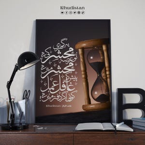 May include: A framed print with white calligraphy and an hourglass. The hourglass has a wooden frame and glass bulbs, with black sand inside. A black desk lamp and other items are on the desk. The print says "Khudistan".