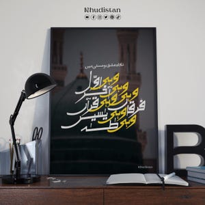 May include: A framed art print with Arabic calligraphy in white and yellow, set against a dark backdrop with a mosque. The print is on a wooden desk, alongside a black desk lamp and an open book.