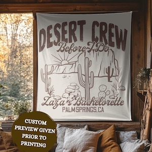 May include: Ivory-colored wall tapestry with the text "DESERT CREW Before I Do" and "Laza's Bachelorette PALM SPRINGS, CA" in brown. The design features cacti and a desert landscape. Available in various sizes, including 26x36 inches.
