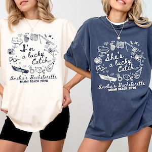 May include: Two t-shirts, one ivory and one denim blue, with a circular graphic design. The design includes the text "I'm a lucky Catch" and "She's a lucky Catch", along with illustrations of a boat, fish, and other elements. The text "Amelia's Bachelorette Miami Beach 2026" is also present.