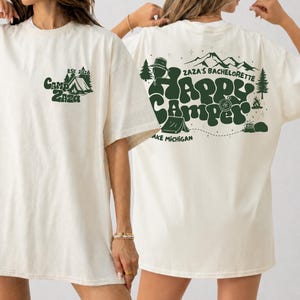 Peut inclure: Un t-shirt ivoire avec des graphismes vert foncé. L'avant présente une tente, des arbres et le texte Camp Zaza Est. 2026. L'arrière affiche le texte Zaza's Bachelorette Happy Camper, des montagnes, des arbres, une tente, une boussole et Lake Michigan.