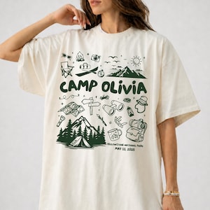 Può includere: T-shirt color avorio con un design grafico verde a tema campeggio e la scritta "CAMP OLIVIA". Il design include illustrazioni di montagne, una tenda e altri elementi all'aperto. È presente anche il testo "YELLOWSTONE NATIONAL PARK MAY 11, 2026".