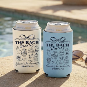 May include: Two can coolers, one ivory and one chambray blue, with the text "THE BACH Diaries" and "Amelia's Bachelorette, Boston, MA." The coolers have a bow design and wedding-themed illustrations. The ivory cooler is on the left.