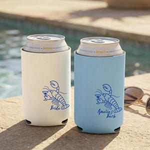 May include: Two cylindrical foam can coolers, one ivory and one chambray blue, each holding a lemon-flavored beverage can. Both feature a royal blue graphic of a lobster holding a martini glass, with the text Bride on one and Amelia's Bach on the other.