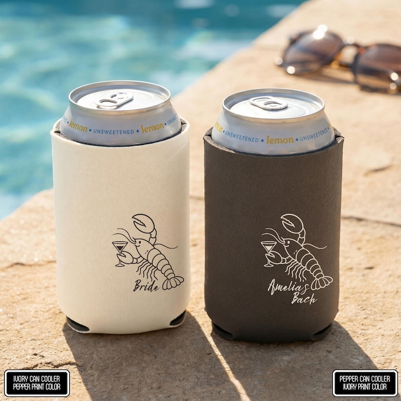 May include: Two foam can coolers, one ivory and one pepper gray, each holding a silver aluminum can labeled lemon unsweetened. The ivory cooler features a black line drawing of a lobster holding a martini glass with the text Bride. The pepper cooler features a white line drawing of a lobster holding a martini glass with the text Amelia's Bach.