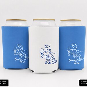 May include: Three foam can coolers, two blue and one white, each featuring a line art illustration of a lobster holding a martini glass. The blue coolers have white text reading Amelia's Bach, while the white cooler has blue text reading Bride.