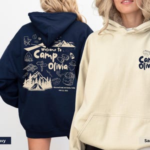 Puede incluir: Una sudadera con capucha azul marino con un gr&aacute;fico color crema en la espalda que muestra monta&ntilde;as, una tienda de campa&ntilde;a y el texto "Welcome to Camp Olivia Yellowstone National Park May 11, 2023" y una sudadera color arena con "Camp Olivia" al frente.