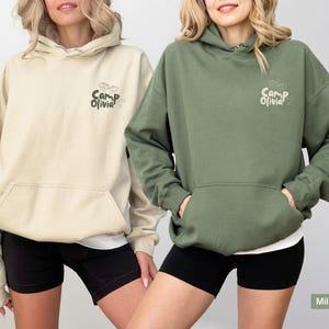 Puede incluir: Dos sudaderas con capucha en colores arena y verde militar. Cada una cuenta con un bolsillo tipo canguro frontal y un peque&ntilde;o gr&aacute;fico en el pecho izquierdo con el texto Camp Olivia junto a una estrella peque&ntilde;a y unos binoculares.