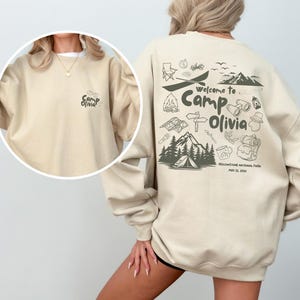 Puede incluir: Una sudadera de cuello redondo color arena con un peque&ntilde;o gr&aacute;fico de 'Camp Olivia' en el pecho izquierdo y un estampado grande en la espalda con iconos de acampada, monta&ntilde;as y el texto 'Welcome to Camp Olivia Yellowstone National Park May 22, 2023'.