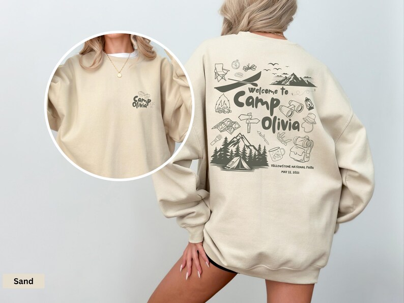 Puede incluir: Una sudadera de cuello redondo color arena con un peque&ntilde;o gr&aacute;fico de 'Camp Olivia' en el pecho izquierdo y un estampado grande en la espalda con iconos de acampada, monta&ntilde;as y el texto 'Welcome to Camp Olivia Yellowstone National Park May 22, 2023'.