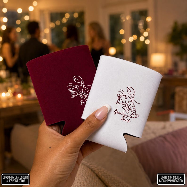May include: Two foam can coolers, one burgundy with a white lobster graphic and one white with a burgundy lobster graphic. The white cooler features the text Amelia's Bach. Both coolers have a curved bottom cutout for a standard beverage can.