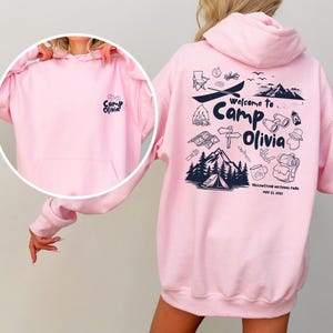 Puede incluir: Una sudadera con capucha rosa claro que presenta un gr&aacute;fico azul oscuro en la espalda con el texto Welcome to Camp Olivia, Yellowstone National Park, May 22, 2021, e iconos de camping. El pecho muestra el texto Camp Olivia.