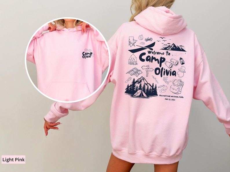 Puede incluir: Una sudadera con capucha rosa claro que presenta un gr&aacute;fico azul oscuro en la espalda con el texto Welcome to Camp Olivia, Yellowstone National Park, May 22, 2021, e iconos de camping. El pecho muestra el texto Camp Olivia.