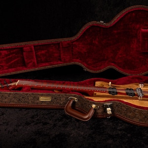 May include: A wooden electric bass guitar rests inside an open, ornate case. The case is brown with a floral pattern and a red velvet interior. The bass guitar has a light wood body with red stripes.