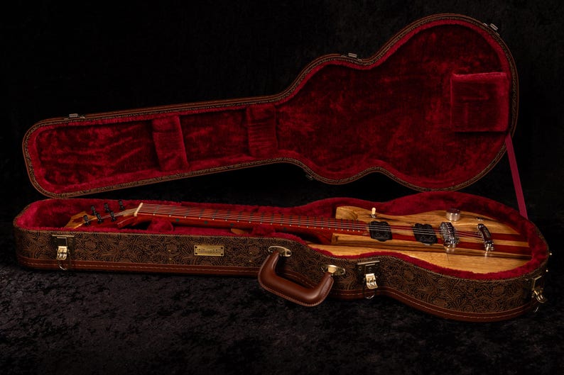 May include: A wooden electric bass guitar rests inside an open, ornate case. The case is brown with a floral pattern and a red velvet interior. The bass guitar has a light wood body with red stripes.