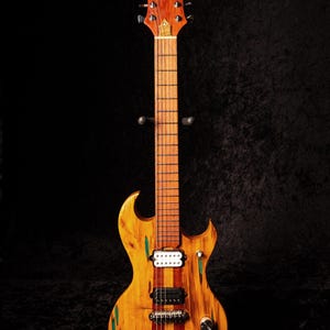 May include: A custom-made electric guitar with a unique wooden body. The guitar features a light brown finish with green and red accents. The headstock has a decorative inlay. The guitar is displayed on a black stand.