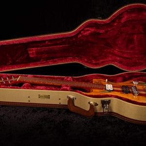 May include: A wooden electric guitar rests inside an open, tweed-covered case with a red velvet interior. The guitar features a striped design with a natural wood finish and chrome hardware. The case has a brown leather handle.