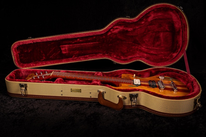 May include: A wooden electric guitar rests inside an open, tweed-covered case with a red velvet interior. The guitar features a striped design with a natural wood finish and chrome hardware. The case has a brown leather handle.